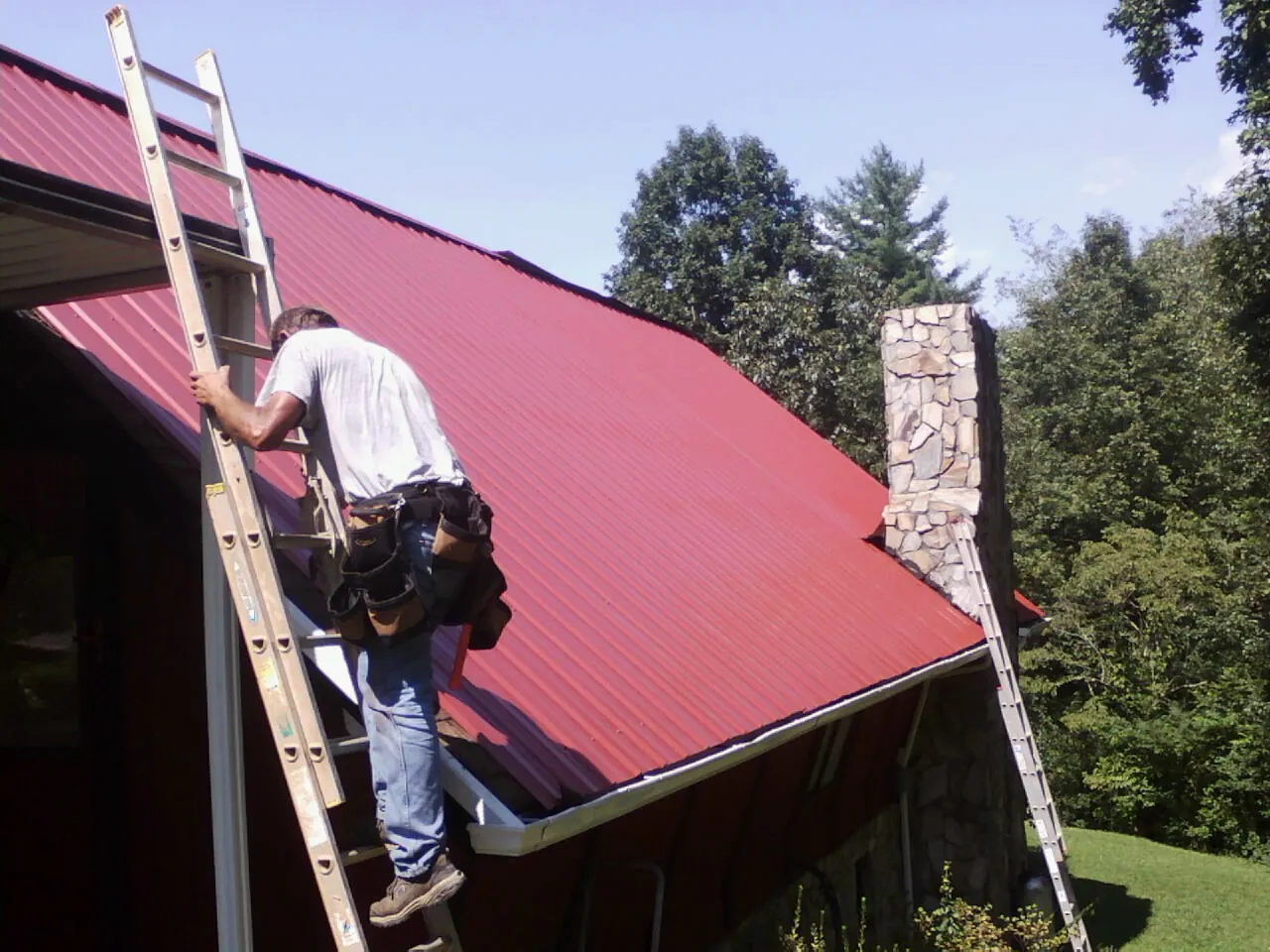 Roofer on a ladder beside a red metal roof and stone chimney — Soffit & Fascia Repair by Roofing Brothers in Huntington Woods